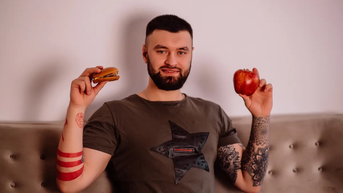 Bearded man pondering diet choice with apple and burger. Indoors with tattoos visible.